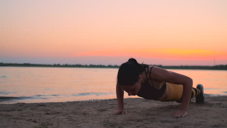 Sporty-girl-doing-push-ups-on-beach-at-sunrise.-Young-fit-tanned-smiling-girl-in-sport-top-and-leggings-doing-push-ups-on-beach-at-sunset